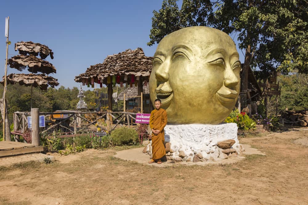 Junger Mönch steht vor einem besonderen Buddha-Kopf - Bambus-Brücke